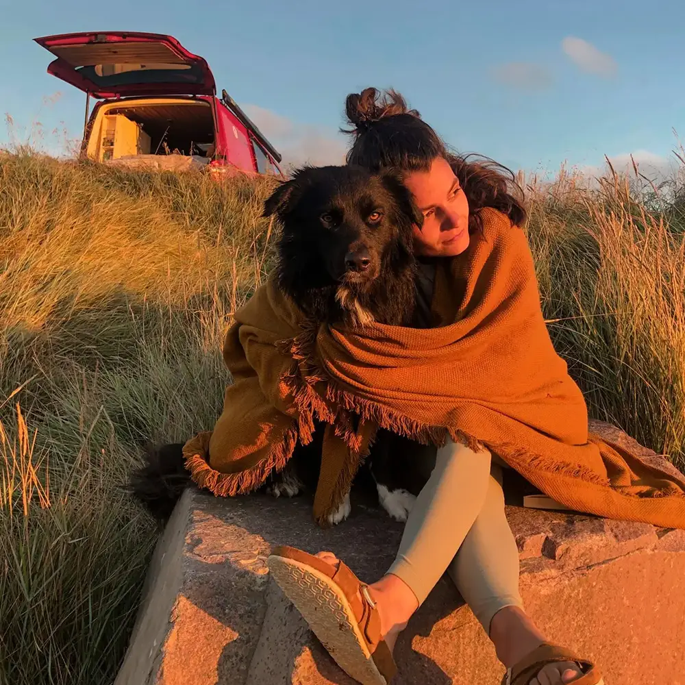 Julia Dillenberg mit ihrem Hund Coffee auf einer Wiese, im Hintergrund ein ausgebauter roter Camper.